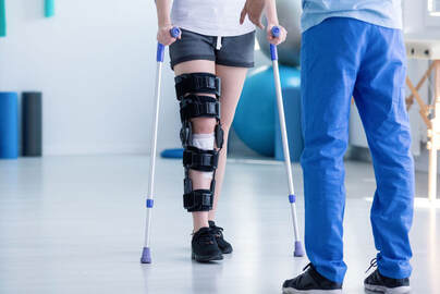 An injured person with a leg brace walks with crutches being assisted by a physical therapist in a rehabilitation room in Lehigh Valley.